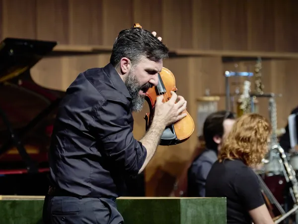 Ein Mann mit dunklem Hemd hält eine Geige an sein Kinn. Im Hintergrund sind Musikinstrumente wie ein Klavier und Holzblasinstrumente sichtbar.