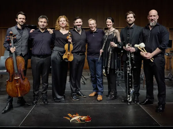 Eine Gruppe von sieben Musikern steht auf einer Bühne. Sie tragen schwarze Kleidung und halten verschiedene Instrumente: ein Cello, eine Violine, eine Klarinette, eine Flöte und ein Percussion-Instrument. Auf dem Boden liegt ein zerbrochenes Musikinstrument.