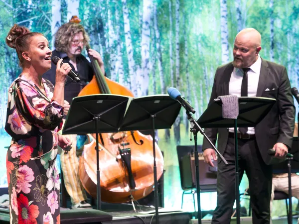 Eine Sängerin in einem bunten, floral gemusterten Kleid steht auf einer Bühne und hält ein Mikrofon. Im Hintergrund ist ein Kontrabassspieler zu sehen. Zwei Mikrofonständer stehen vor den Musikern.