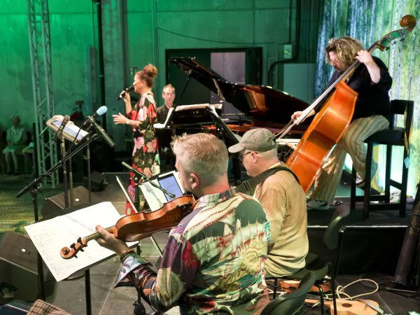 Eine Musikergruppe auf einer Bühne. Eine Sängerin mit Mikrofon steht vorne, während ein Pianist, ein Kontrabassist und ein Geiger im Hintergrund spielen. Die Musiker tragen bunte Kleidung.