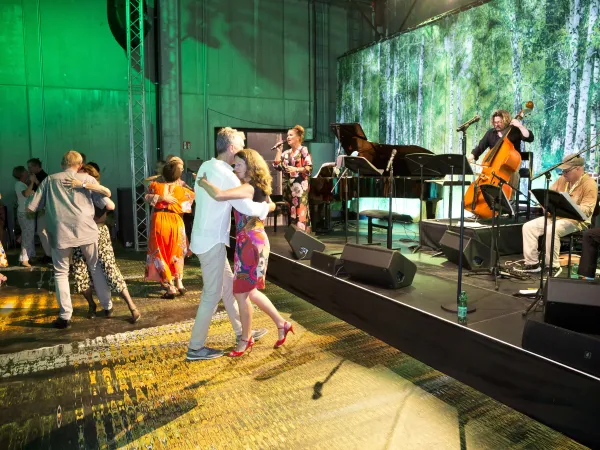 Tanzende Paare auf einer Bühne, umgeben von einer grünen Waldkulisse. Im Hintergrund eine Band mit Klavier, Kontrabass und Mikrofonen.