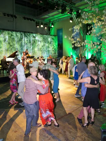 Tanzende Paare auf einer Veranstaltung, umgeben von einer grünen, baumartigen Kulisse. Im Hintergrund spielen Musiker an einem Klavier.