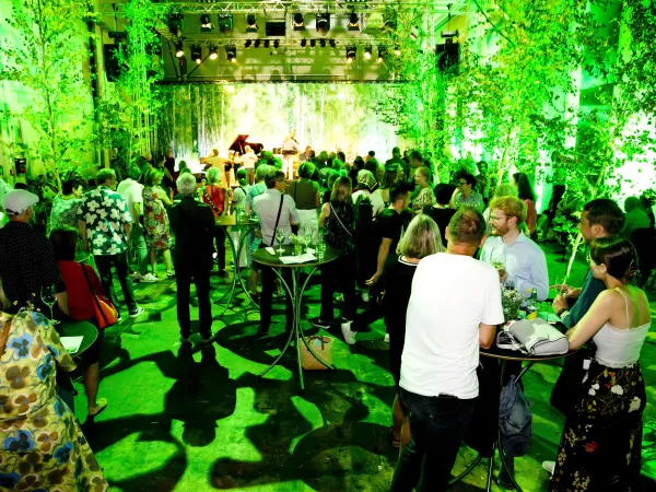 Eine große Menschenmenge steht in einem Raum mit grüner Dekoration und Pflanzen. Im Hintergrund sind zwei Musiker auf einer Bühne sichtbar. Tische stehen verteilt im Raum.