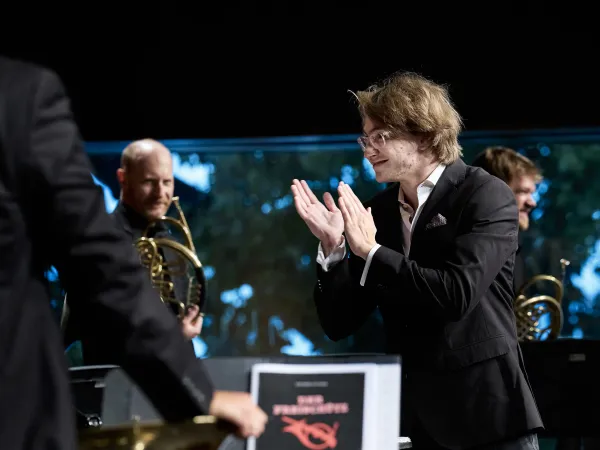 Ein Mann in einem schwarzen Anzug mit einem weißen Hemd applaudiert. Im Hintergrund sind Musiker mit Hörnern zu sehen. Auf einem Notenständer steht 'DAS PHILHARMONIKER'.