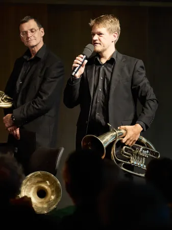 Ein Musiker in schwarzem Anzug hält ein Mikrofon in der rechten Hand und eine Tuba in der linken Hand. Im Hintergrund steht ein weiterer Musiker mit einem Horn.