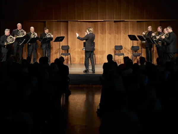 Ein Orchester mit sieben Hornspielern in schwarzer Kleidung steht auf einer Bühne. Ein Dirigent in einem schwarzen Anzug leitet sie. Im Hintergrund sind Holzpaneele sichtbar.