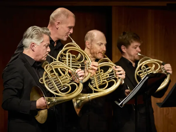 Fünf Musiker in schwarzer Kleidung spielen Hörner. Der erste Musiker hält ein großes, gebogenes Horn, während die anderen drei ihre Instrumente in der Hand halten.