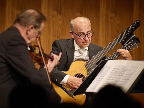 Zwei Musiker auf einer Bühne. Der Mann links spielt eine Violine, der Mann rechts spielt eine Gitarre mit sechs Saiten. Notenblätter sind auf einem Ständer sichtbar.
