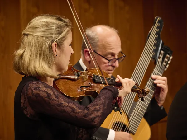 Eine Frau mit blonden Haaren spielt eine Geige. Im Hintergrund steht ein Mann mit einer Gitarre und einem großen Saiteninstrument.