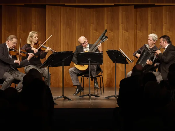 Fünf Musiker auf einer Bühne, die verschiedene Instrumente spielen: zwei Geigen, ein Akkordeon und eine Gitarre. Sie tragen formelle Kleidung und stehen um Notenständer.