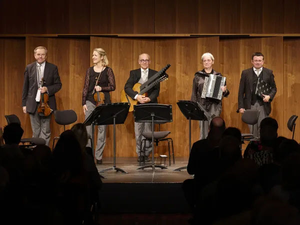 Fünf Musiker auf einer Bühne, zwei mit Streichinstrumenten, einer mit Gitarre, einer mit Akkordeon und einer mit einem anderen Instrument. Alle tragen formelle Kleidung.
