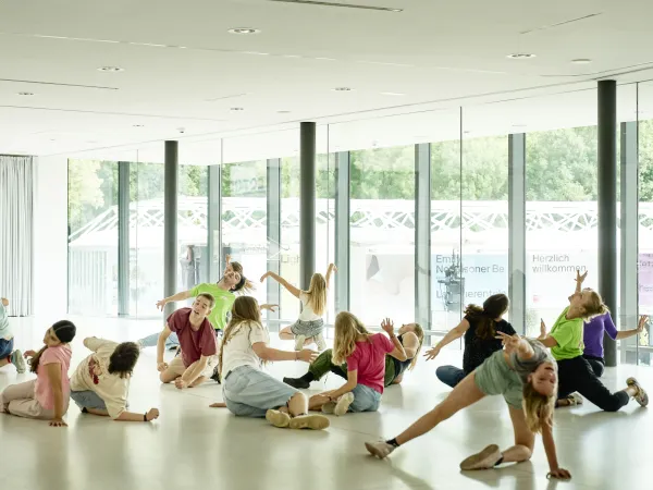 Gruppe von Kindern und Jugendlichen in einem Tanzraum, die verschiedene Posen einnehmen. Einige liegen auf dem Boden, andere strecken die Arme aus. Im Hintergrund sind große Fenster mit Blick ins Freie.