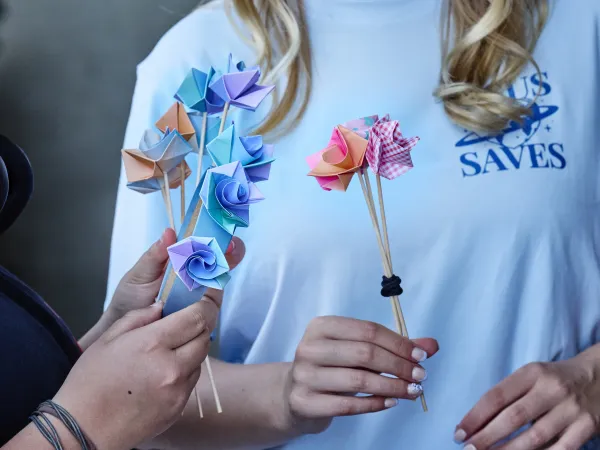 Zwei Personen halten bunte Origami-Rosen aus Papier in verschiedenen Farben und Mustern. Eine Person hat eine blaue und eine orange Rose, die andere eine rote und eine karierte Rose. Beide tragen T-Shirts.
