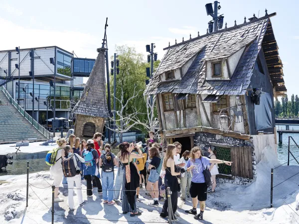 Gruppe junger Menschen steht vor einem großen, alten, schiefen Haus mit einem steinernen Kamin und einem Baum. Im Hintergrund sind moderne Gebäude und Lichtanlagen sichtbar.