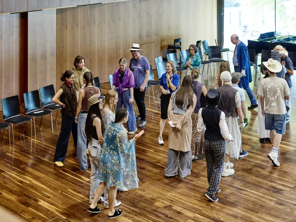 Eine Gruppe von jungen Menschen in verschiedenen Outfits interagiert in einem Raum mit Holzboden. Einige tragen Hüte und Accessoires, während andere in lässiger Kleidung gekleidet sind. Stühle stehen im Hintergrund.