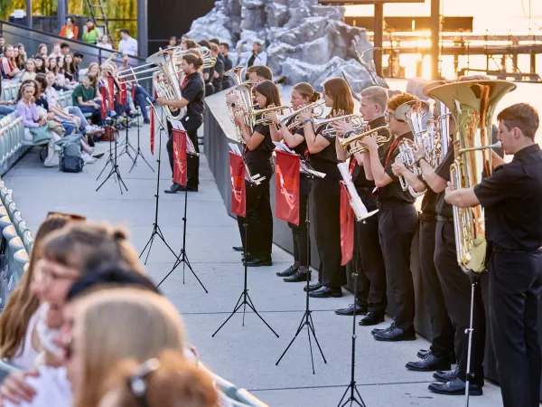 Eine Gruppe von Musikern in schwarzer Kleidung spielt Blechblasinstrumente auf einer Freiluftbühne. Im Hintergrund sitzen Zuschauer auf einer Tribüne.