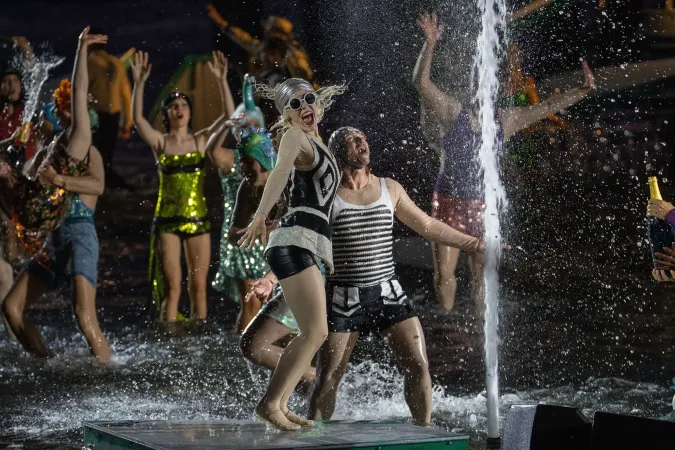Menschen in bunten Kostümen tanzen und springen um eine Wasserfontäne auf einer Bühne