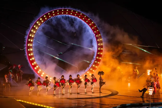Tänzerinnen in roten Kostümen mit schwarzen Federkopfschmuck führen eine Choreografie vor einem großen, beleuchteten Ring mit Nebel auf einer Bühne auf