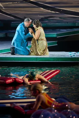 Ein Mann im blauen Anzug und eine Frau im goldenen Kleid knien sich auf einer Bühne über Wasser und halten sich an den Händen, im Vordergrund liegen zwei Personen auf roten Luftmatratzen im Wasser.