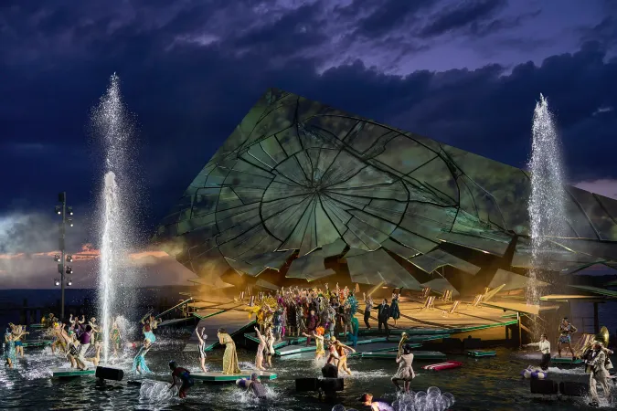 Bühnenbild mit großem schräg gestelltem, zerbrochen wirkendem Rad im Hintergrund und zahlreichen Schauspielern auf einer Wasserbühne mit zwei hohen Wasserfontänen bei Dämmerung