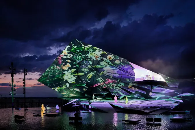 Bühnenaufbau mit farbiger Projektion auf einer Wasserbühne unter bewölktem Abendhimmel mit mehreren Personen auf der Bühne und im Wasser