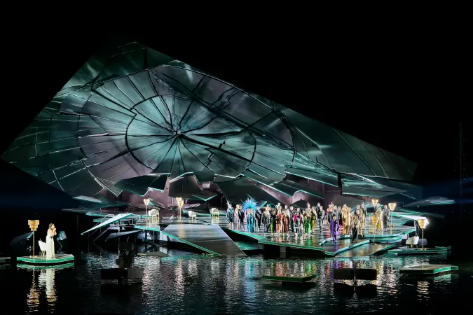 Bühnenbild mit großer, zerbrochener Uhr als Kulisse und Ensemble auf einer Wasserbühne bei einer Opernaufführung